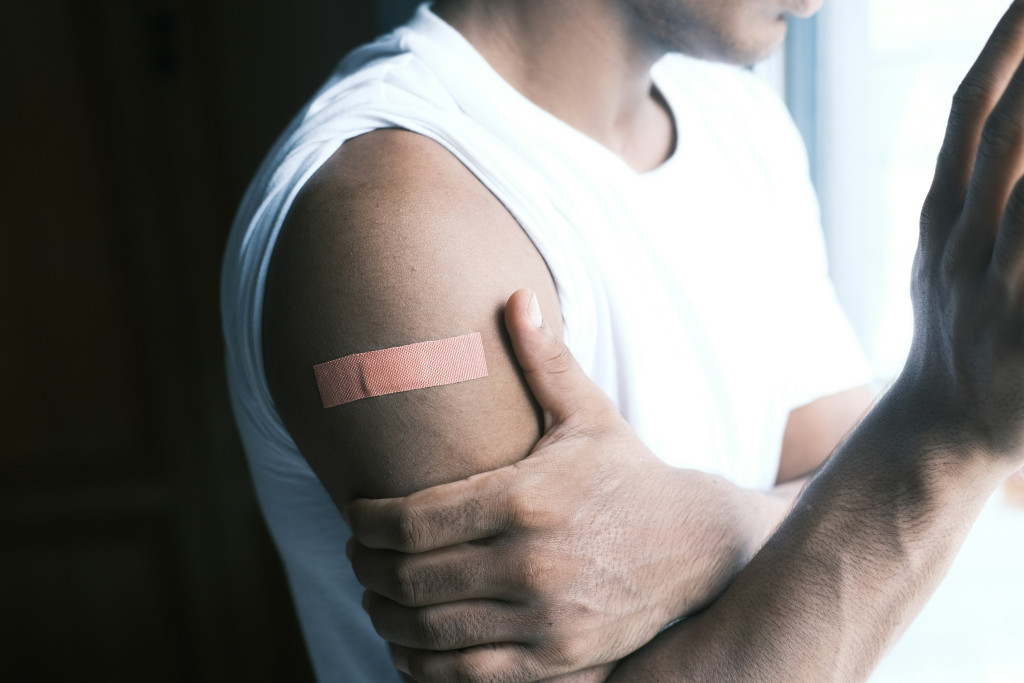man in a white tank top holding his bicep with a post-injection band aid on it
